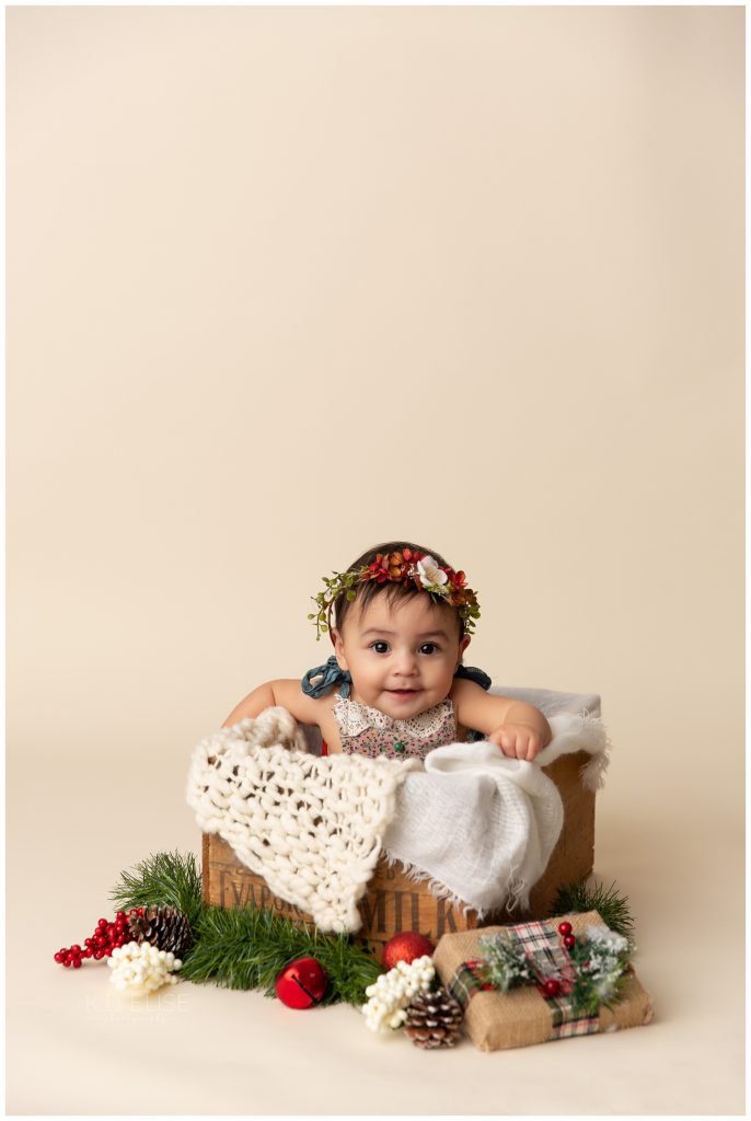 Baby girl in a Christmas out and floral headband sitting in a wooden crate. Six month milestone session by K.D. Elise Photography.