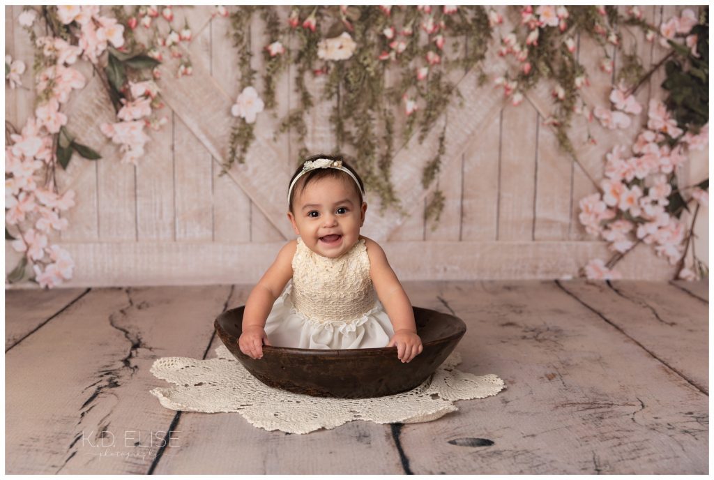 Baby girl sitting in a wooden bowl in front of a wood and floral backdrop.