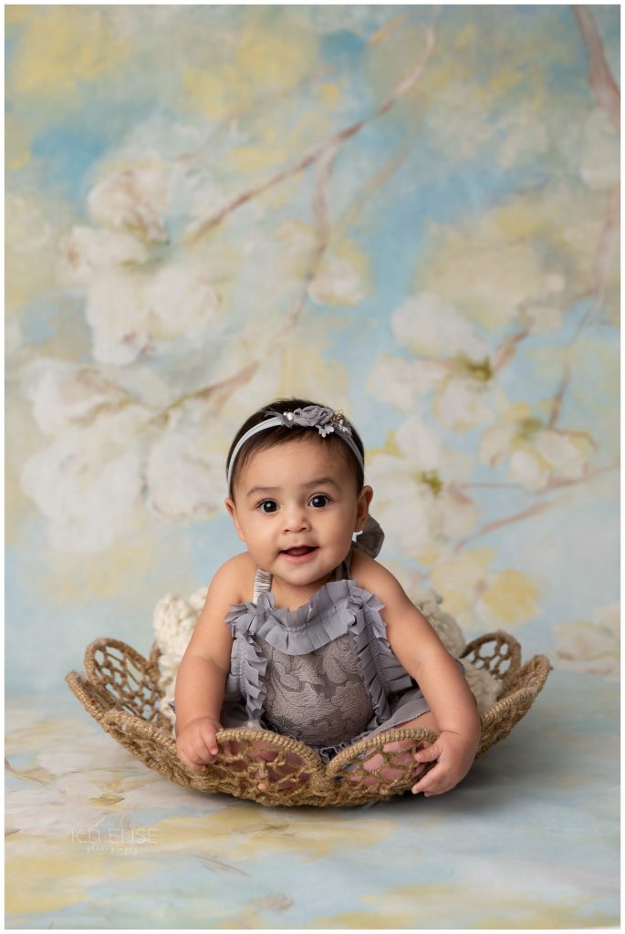 Baby girl in grey romper, sitting in a basket in front of floral backdrop. Sitter milestone session from K.D. Elise Photography.