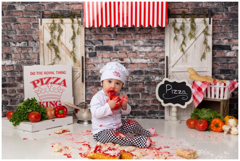 Pizza themed cake smash photo of little boy in chef hat and pants. He's sitting in front of a brick backdrop, under a red awning. He's holding a tomato, while covered in red frosting and surrounded by cake mess.