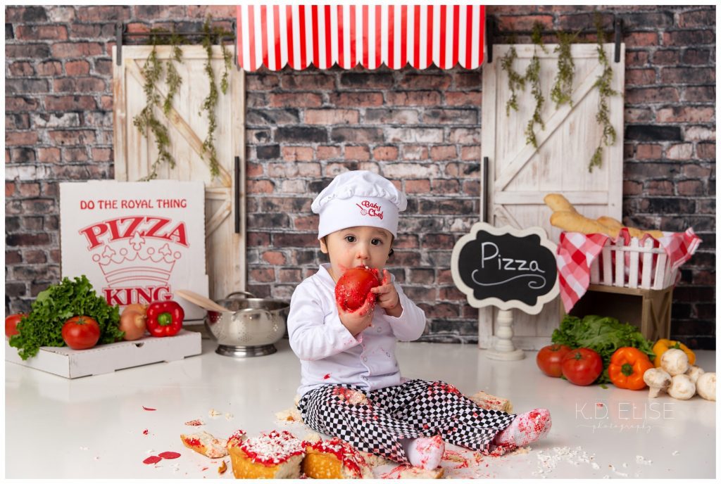 Pizza themed cake smash photo of a baby boy in a chef costume, eating a tomato. He is sitting beside a pile of broken up cake. The backdrop is brick, with a red and white striped awning and a white wooden door on either side. 