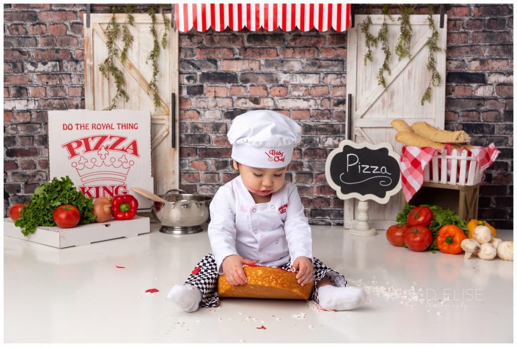 Pizza themed cake smash photo of a baby boy in a chef costume. He's in front of a brick backdrop, with a red awning and white doors. He's trying to lift a cake that looks like a deep dish pizza.