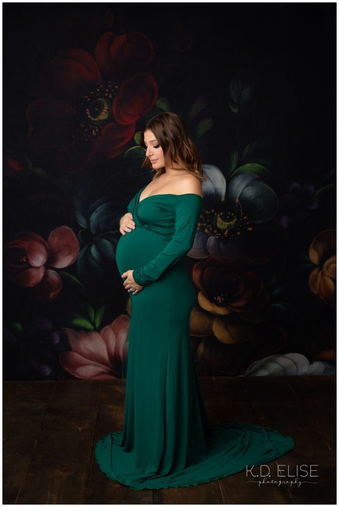 In studio maternity photo of pregnant mom in a green gown, standing in front of a black floral backdrop.