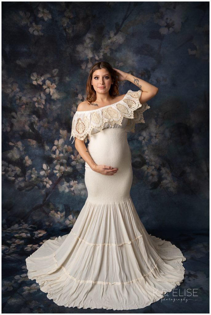 In studio maternity session, photo of pregnant mom in a cream mermaid style gown. She's standing in front of a blue and cream floral backdrop.