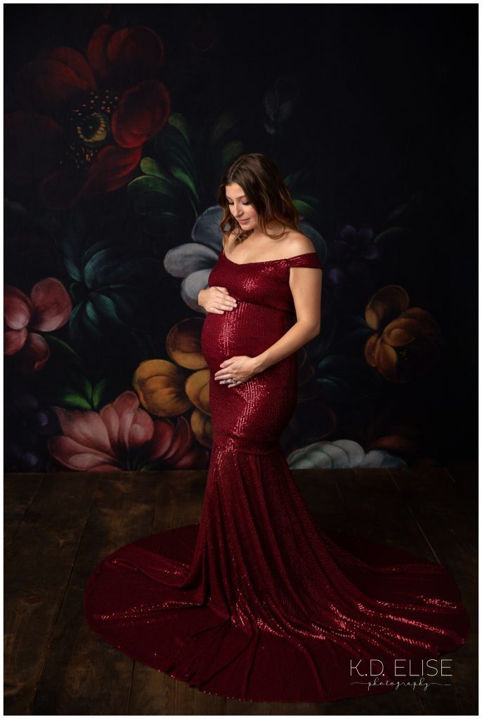 In studio maternity session photo of a pregnant mom in a red sequin dress. She's holding and looking down at her belly. The backdrop is black, with large colorful flowers. 