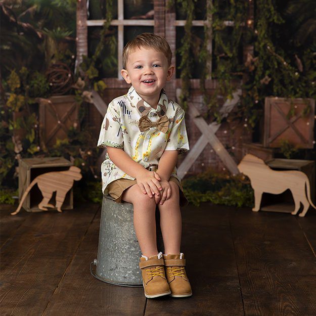 Little boy in khakis, a white shirt, and a bowtie smiles for the camera while sitting on a metal bucket.