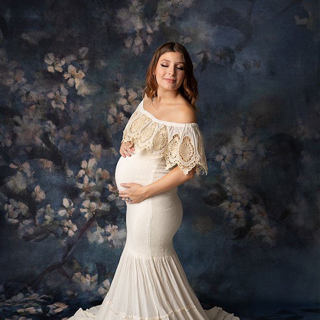 Pregnant mom in a white lace gown poses for maternity photos in front of blue floral backdrop.